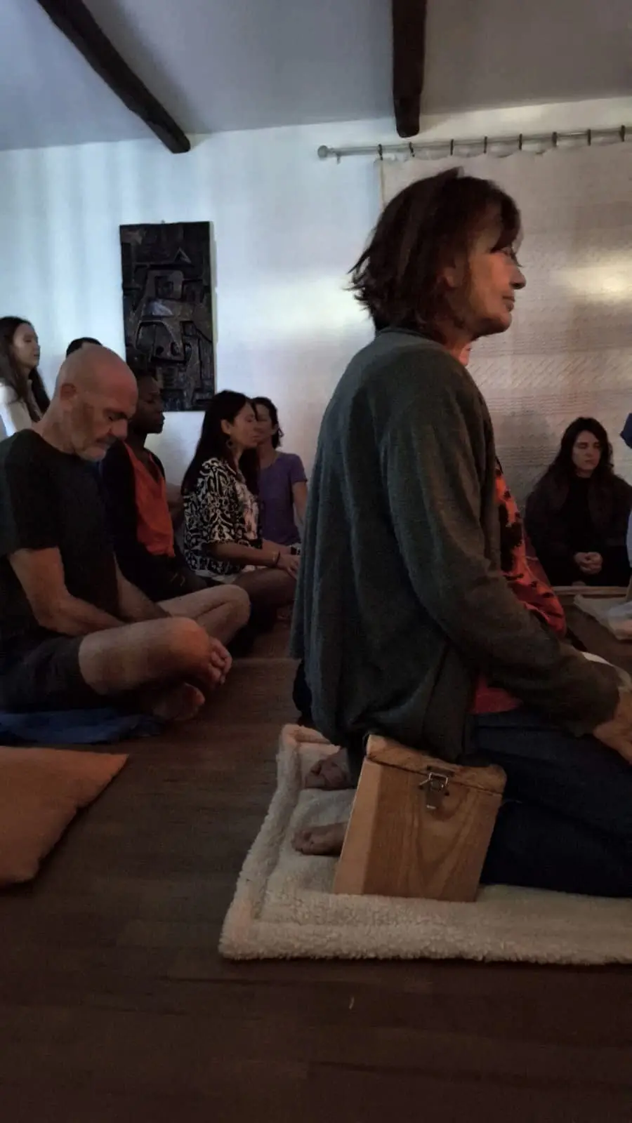 Durante una meditazione di gruppo: in primo piano una donna in ginocchio sulla panchetta di legno, dietro un uomo a gambe incrociate sul cuscino